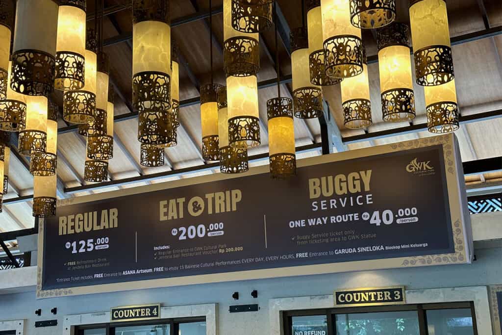 Hanging lanterns glow above a sign listing prices for regular, eat & trip, and buggy services at Garuda Wisnu Kencana.