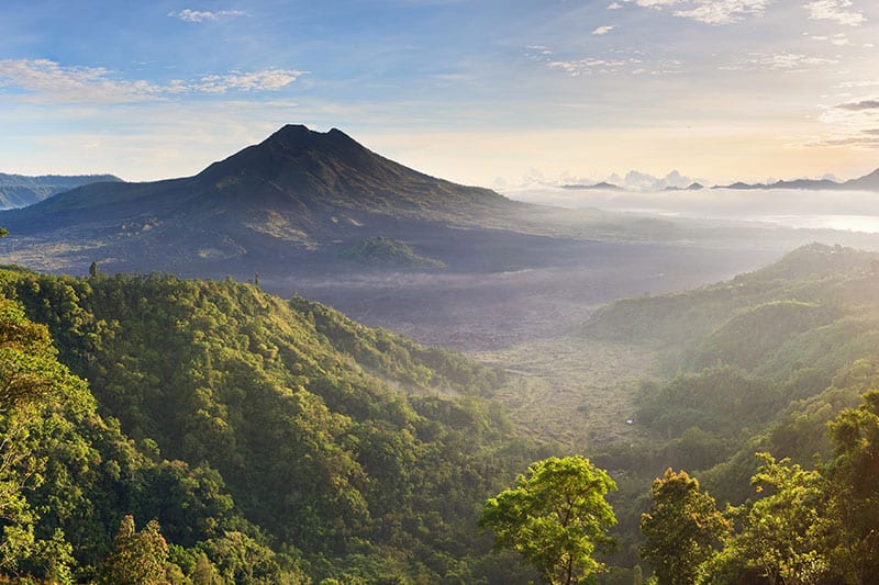 A mountain landscape in Bali showcases lush green hills and a prominent volcanic peak in Kintamani, all under a clear sky at sunrise.