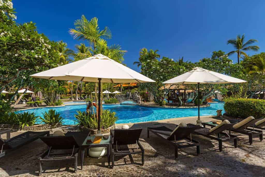 A tropical resort pool area in Bali features wicker lounge chairs under white umbrellas, surrounded by lush green trees and plants. The vivid blue pool mirrors the clear sky, with palm trees swaying gently in the background.