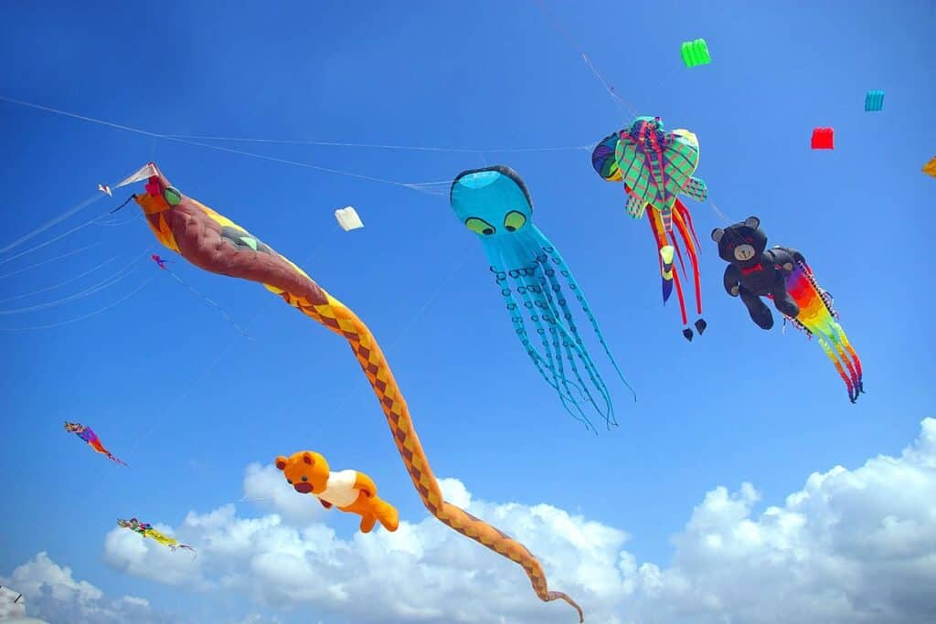 Colorful kites in various shapes, including an octopus, snake, and teddy bear, dance above Bali’s clear blue sky with fluffy white clouds.