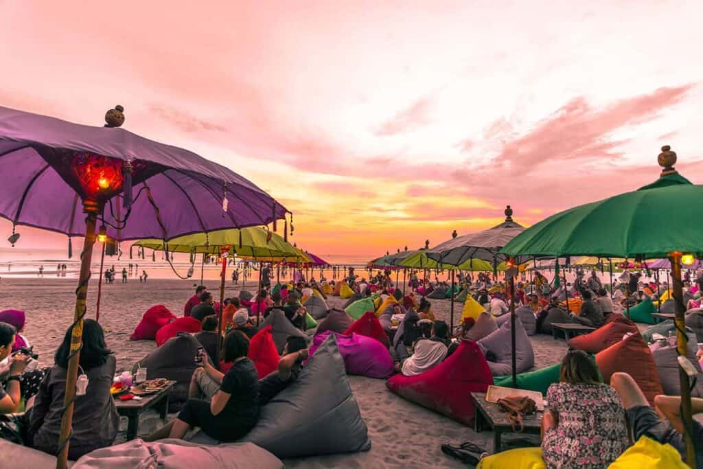 At Seminyak's Double Six Beach, a vibrant sunset casts a warm glow over a sea of colorful umbrellas and comfy bean bags, where crowds gather to relish the breathtaking view.