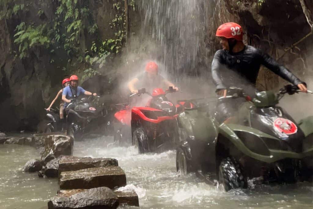 Riders navigate their ATVs through Bali's misty waterfall and rocky stream, donning helmets and protective gear.