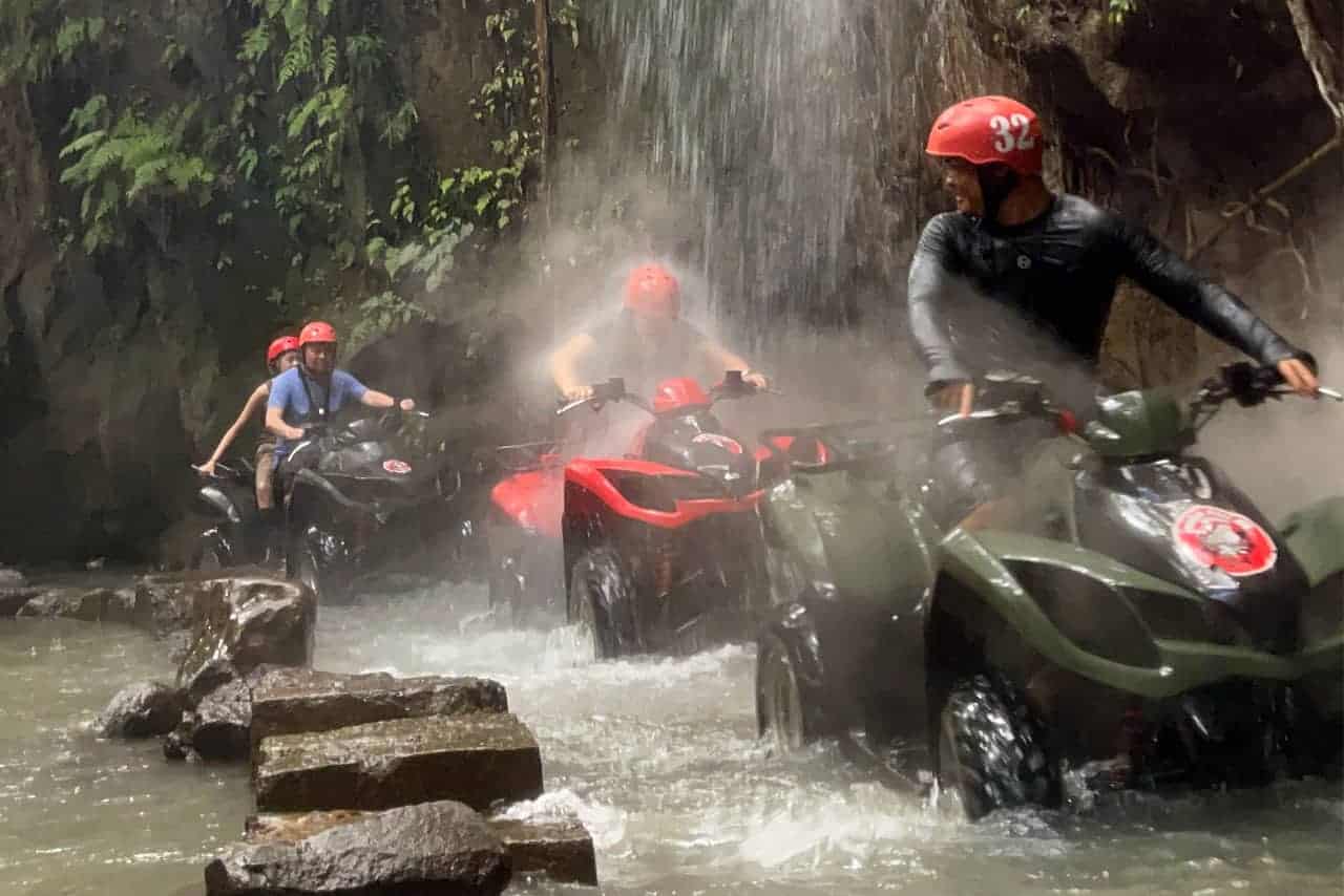 Riders navigate their ATVs through Bali's misty waterfall and rocky stream, donning helmets and protective gear.