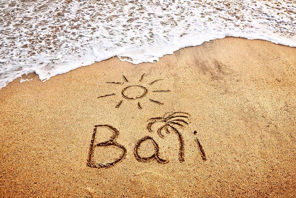 The word Bali is written in sand with sun and palm trees, where ocean waves lovingly embrace the shore.