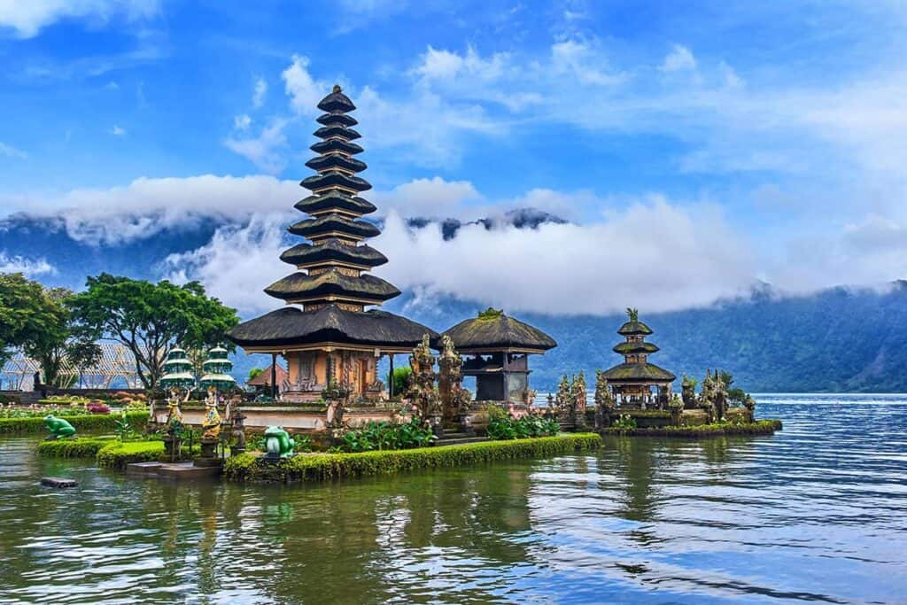 Nestled in Bali, a serene lakeside temple with multi-tiered pagoda roofs is surrounded by lush greenery and mountains under a cloudy sky. The reflection of the temple and clouds dances gracefully in the shimmering water.