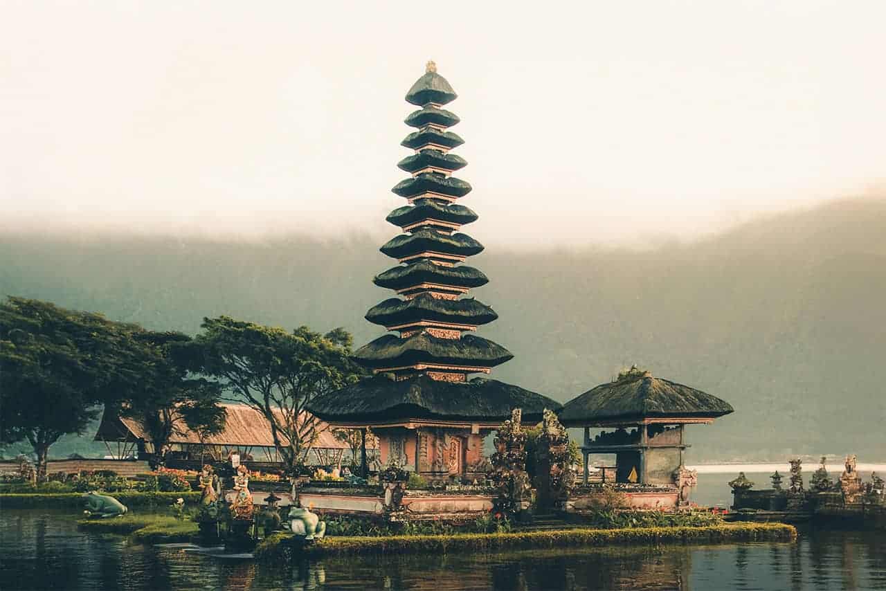 Ancient temple with a tiered roof rests on the serene waters of Lake Beratan in Bali, surrounded by lush trees and misty mountains.