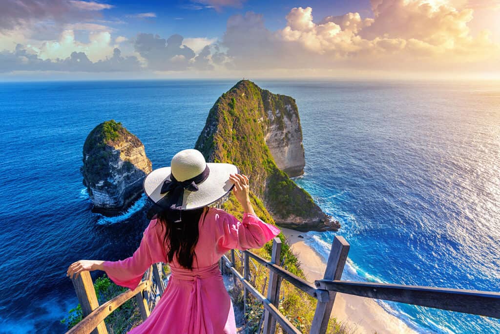 Woman in a pink dress and hat gazes at the dramatic ocean cliffs and sands of Kelingking Beach from a high viewpoint at sunset.