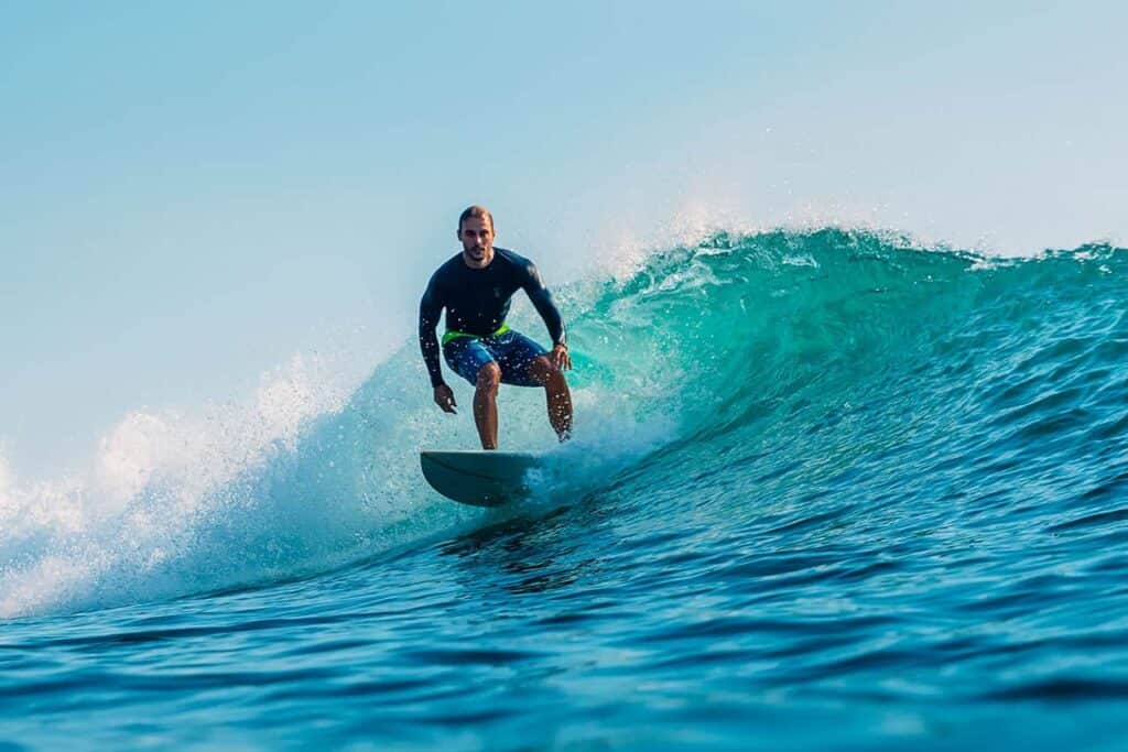 A surfer wearing a black rash guard and colorful shorts rides a turquoise wave under the clear Bali sky. The wave curls behind him as he skillfully balances on his surfboard, capturing the essence of this tropical paradise.