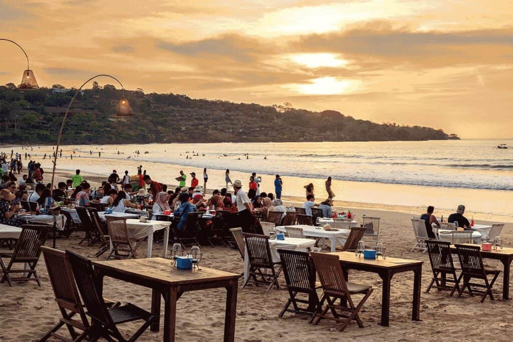 Nestled in the picturesque setting of Jimbaran, this Bali beachside restaurant offers diners a stunning sunset view over the ocean, framed by empty tables in the foreground, inviting more guests to join the seaside experience.