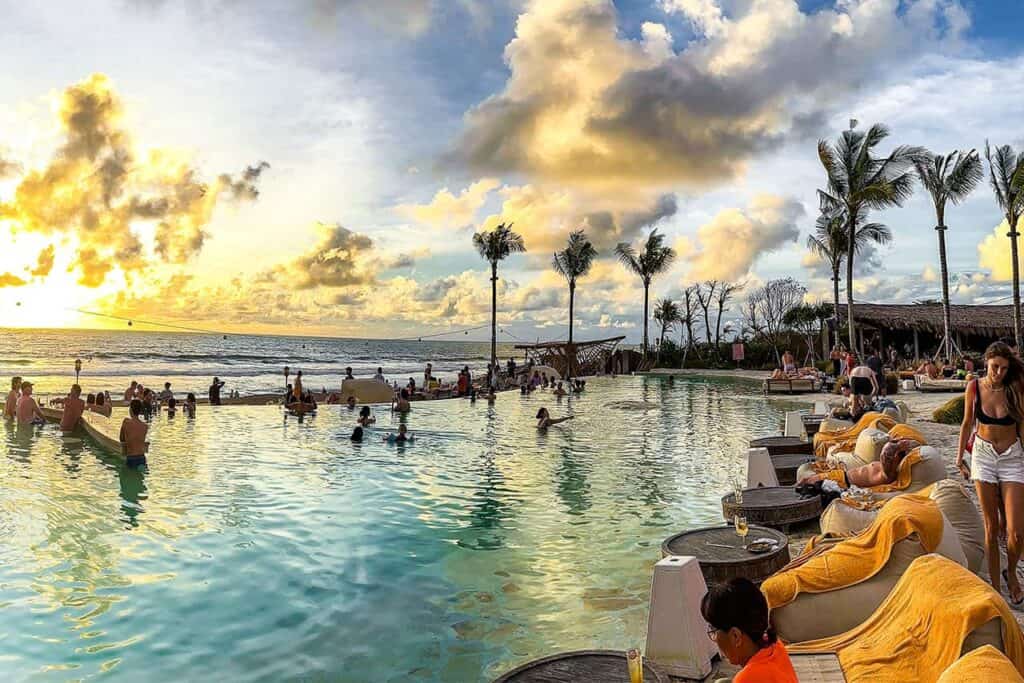 People relax by a large beachside pool at sunset in Kuta, with Bali's palm trees and golden skies painting the background.