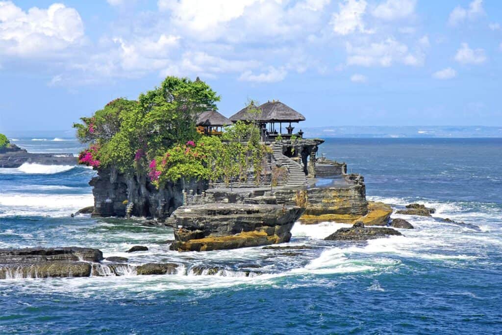 The ocean temple on this rocky island, reminiscent of Bali's Tanah Lot, is adorned with green foliage and pink flowers, all embraced by waves under a serene blue sky.