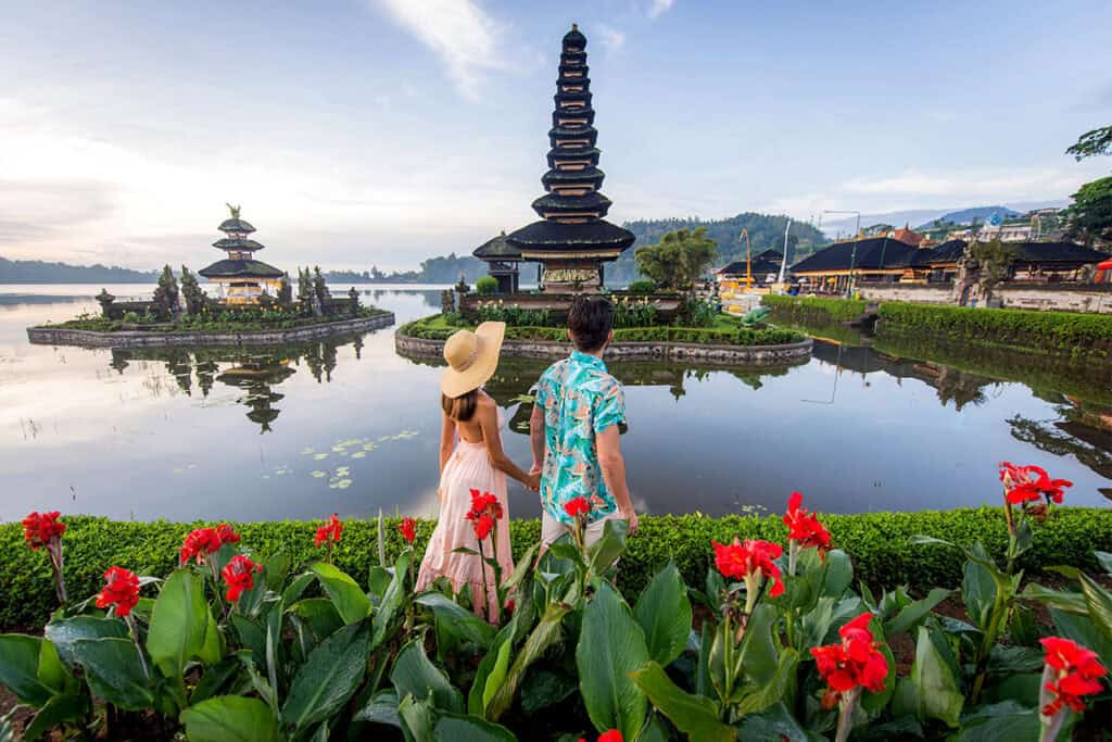 A couple stands by Lake Danu Beratan, facing a Balinese temple surrounded by red flowers and lush greenery.