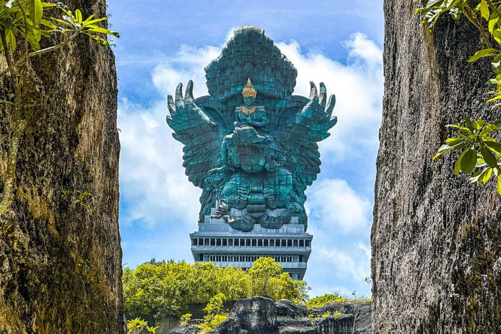 The majestic Garuda Wisnu Kencana statue rises between rocky cliffs and lush greenery, set beneath a blue sky dotted with clouds.