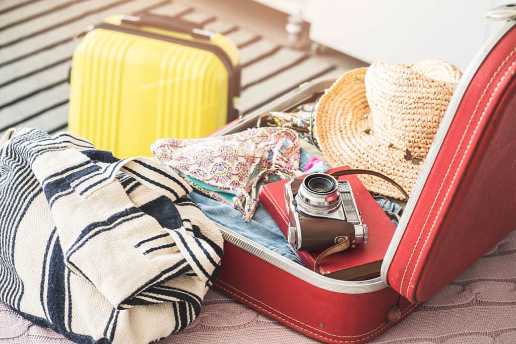 Open red travel suitcase with clothes, straw hat, and camera; yellow suitcase in background on striped floor.