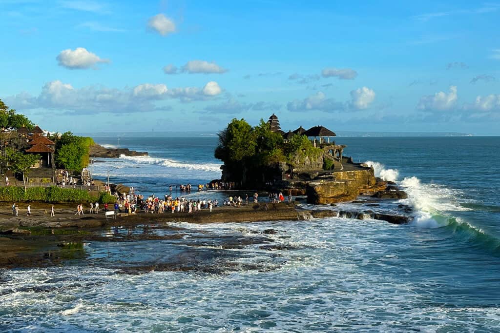Crowds gather on the rocky shore near Tanah Lot Temple, with waves crashing under a bright, blue sky.