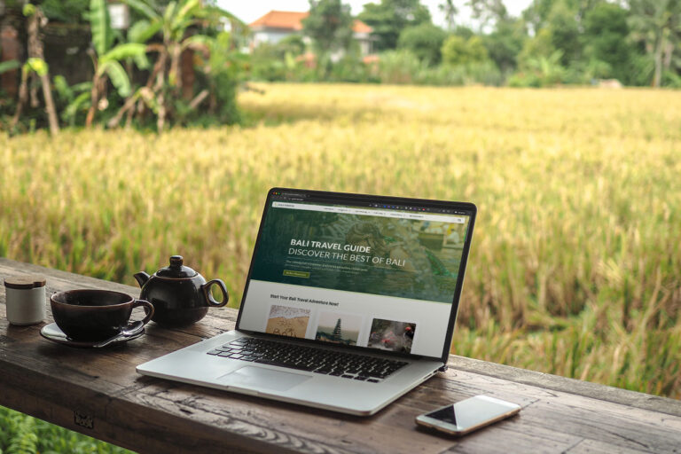 Laptop, tea set, and phone on a wooden table overlooking a rice field and trees in a rural setting.